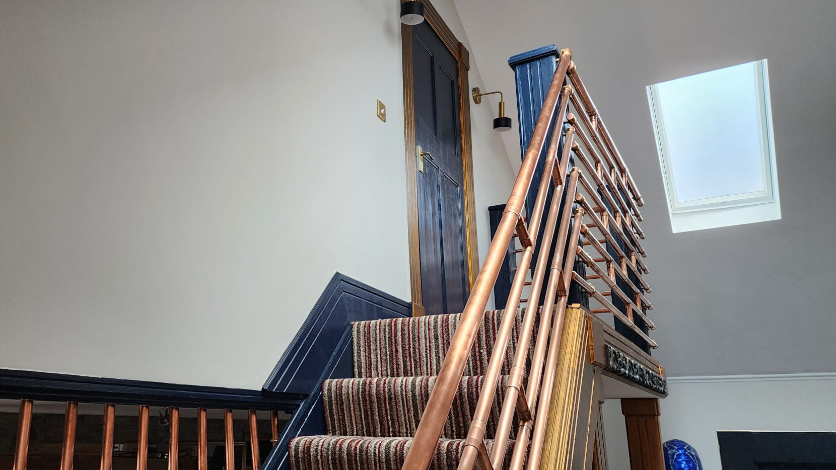Blue skirting board with a staircase covered in lined coloured carpet with copper handrails, with skylight to let natural light in.