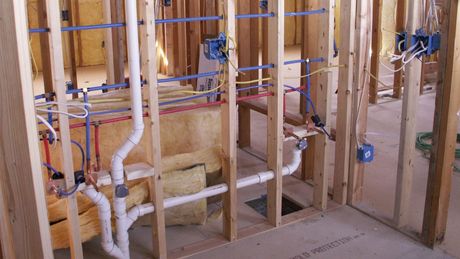 Exposed wall framing in a house under construction, showing plumbing pipes, electrical wiring, and insulation between wooden studs.
